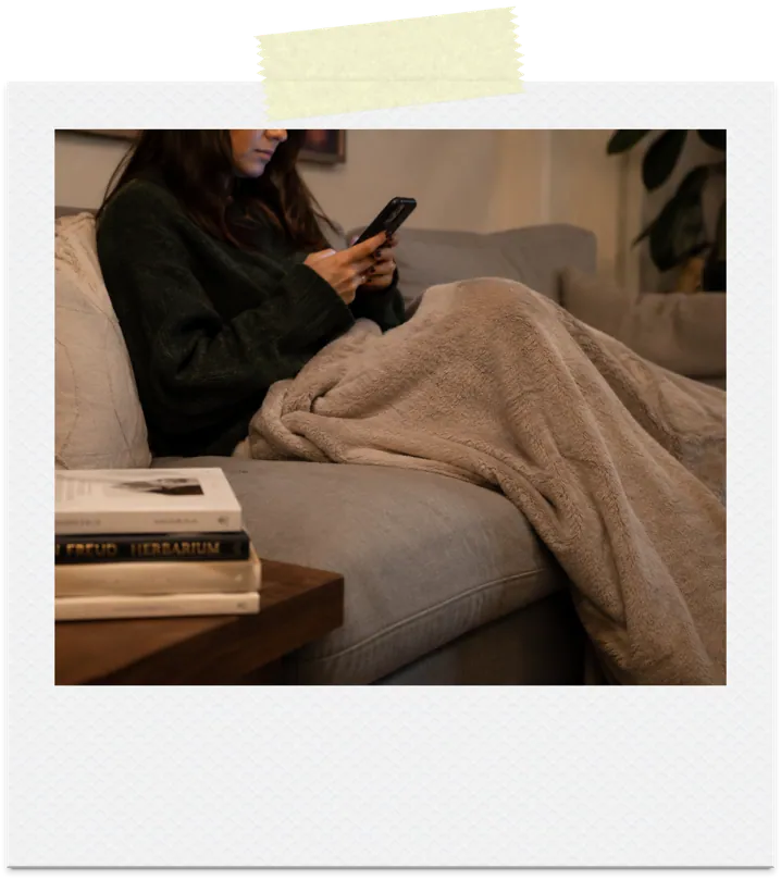 A woman scrolling her phone under a blanket instead of reading the books beside her